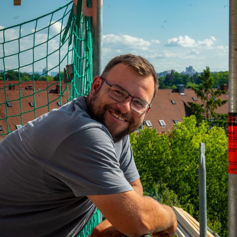Lächelnder Dachdecker im Schäfer-Shirt am Gerüst, Ortspanorama im Hintergrund